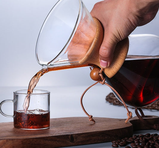 Chemex Coffee Maker pouring coffee into a glass cup on a wooden surface with coffee beans in the background.