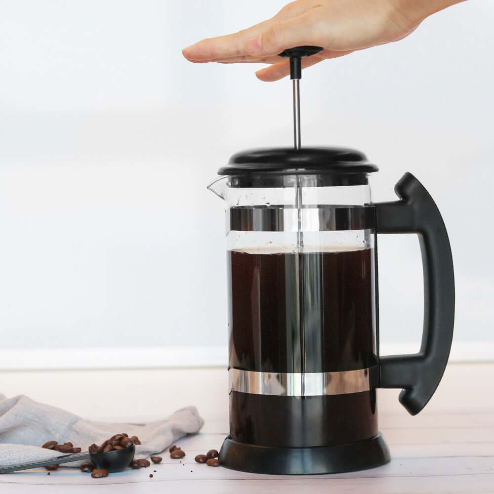 Hand pressing down on the plunger of a Glass French Press to brew coffee.