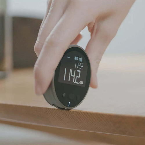 A hand holds the PrecisionCore HD, a High Precision Electronic Measuring Ruler Tool, showing a 114.2 reading on its black circular digital screen against wood—ideal for accurate measurements.