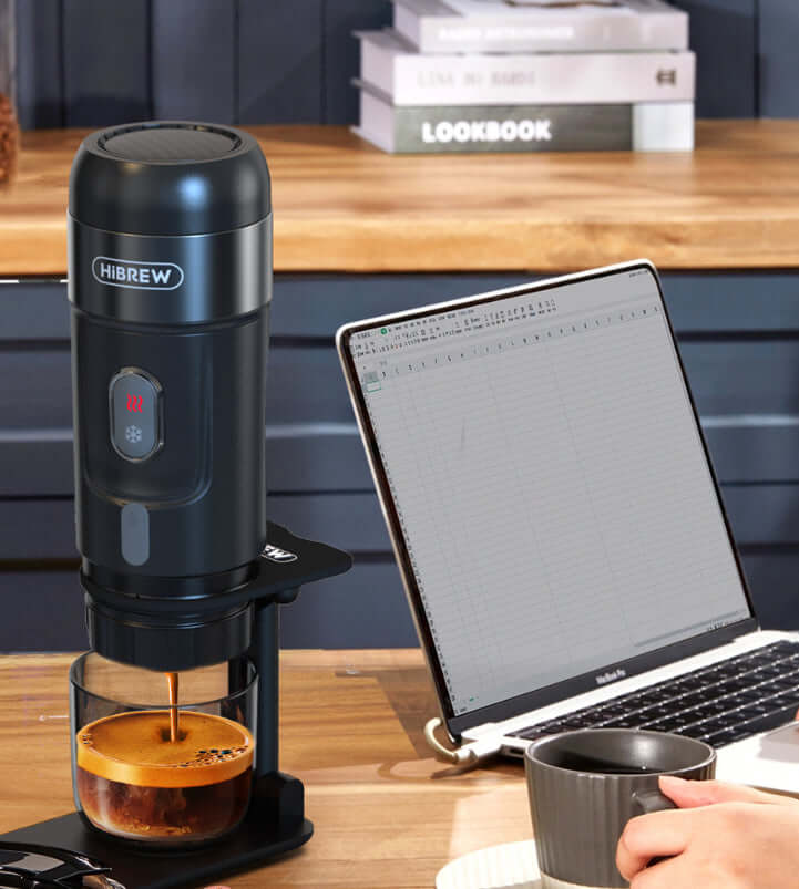Portable Electric Espresso Maker brewing coffee next to a laptop on a wooden table.