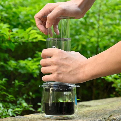 Person using a Portable Espresso Maker in a green outdoor setting, showcasing its hand-pressure brewing method.