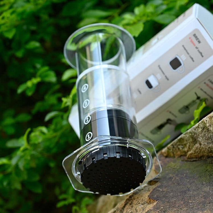 Portable Espresso Maker next to packaging on rock with green foliage in the background.